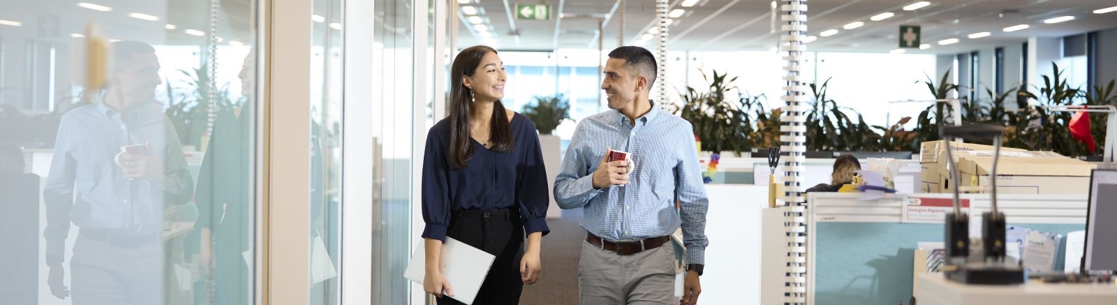 A woman and man walking in an office. They are smiling and looking at each other. The man is holding a coffee cup and the woman is holding a folder
